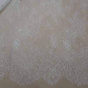 Pode incluir: Tecido de renda floral branca com borda ondulada. A renda apresenta um delicado padr&atilde;o floral e est&aacute; parcialmente enrolada. Uma etiqueta com o texto "Lace Fabrics" &eacute; vis&iacute;vel.