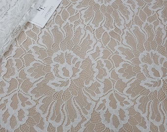 May include: A white lace fabric featuring a large-scale floral pattern with intricate scalloped edges. A small white rectangular tag with the black text "LACE FABRICS" is visible in the upper left corner, partially covered by a folded section of the lace.