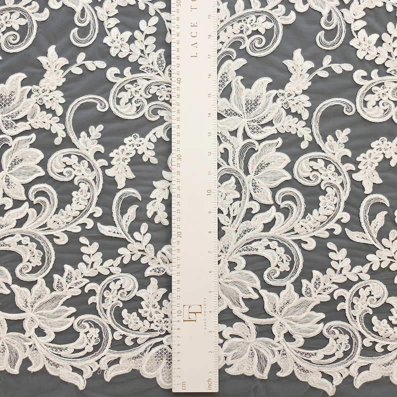May include: Off-white lace fabric featuring an intricate floral and scroll pattern with embroidered details. A white ruler is placed vertically over the fabric, showing measurements in both inches and centimeters for scale reference.