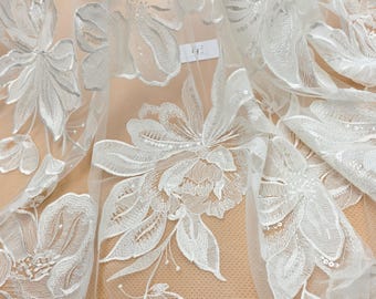 May include: Close-up of white floral lace fabric. The fabric features detailed embroidered flowers and leaves with sequin accents. The delicate design is set against a sheer background, creating a textured and elegant look. Ideal for bridal or formal wear.