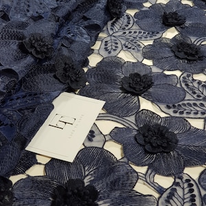 May include: A navy blue floral lace fabric featuring three-dimensional flower appliqu&eacute;s with intricate embroidery and cut-out details. A white rectangular tag is placed on the fabric with the logo LT and the text LACE TO LOVE printed in black.