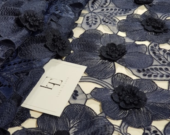 May include: A navy blue floral lace fabric featuring three-dimensional flower appliqués with intricate embroidery and cut-out details. A white rectangular tag is placed on the fabric with the logo LT and the text LACE TO LOVE printed in black.