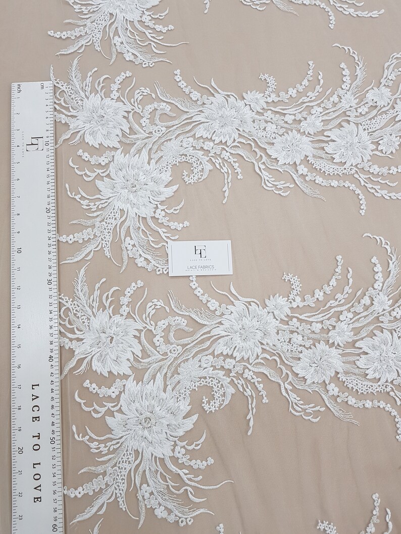 Puede incluir: Tela de encaje floral blanca con un delicado patr&oacute;n de flores y hojas. El encaje est&aacute; sobre un fondo de malla transparente.