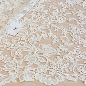 May include: White lace fabric featuring an intricate floral and vine embroidery pattern with scalloped edges. The lace is layered over a solid beige background and includes a small white rectangular tag with a black logo in the upper left corner.