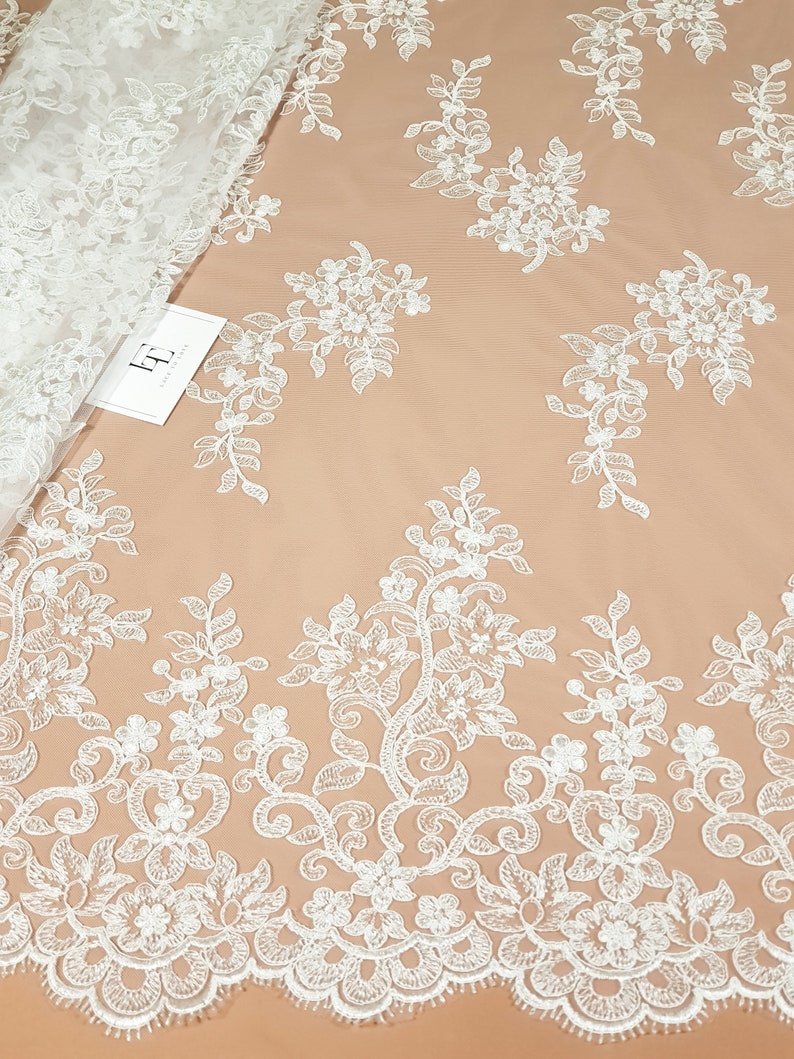 May include: White floral lace fabric with a delicate pattern of flowers and leaves. The lace has a scalloped edge and is perfect for adding a touch of elegance to any project.