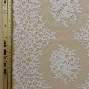 May include: White lace fabric with a floral pattern. The lace is delicate and intricate, with a scalloped edge. The fabric is shown against a beige background.