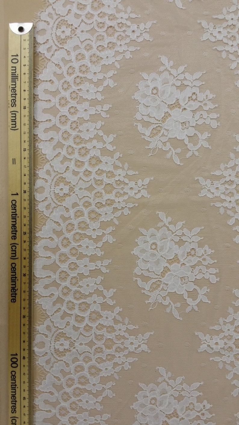 May include: White lace fabric with a floral pattern. The lace is delicate and intricate, with a scalloped edge. The fabric is shown against a beige background.