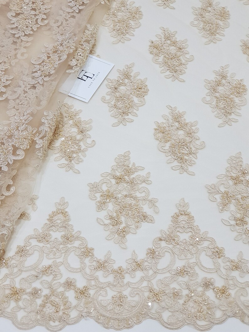 May include: Beige lace fabric with floral embroidery and beaded accents. The fabric has a repeating pattern of embroidered flowers and a scalloped edge. A tag with the text "Lace to Love" is visible.