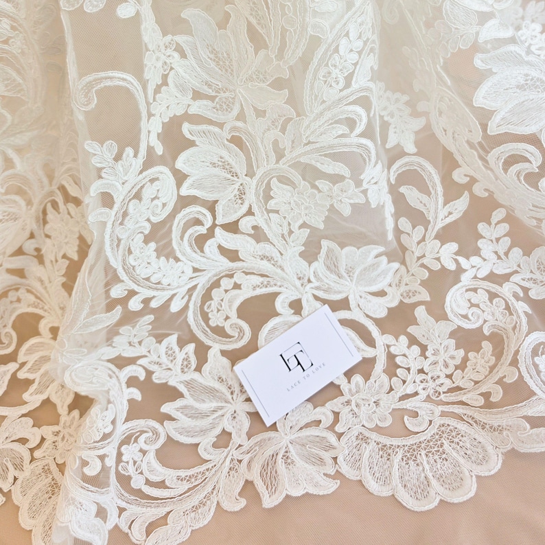 May include: Off-white lace fabric featuring an intricate floral and vine embroidery pattern on a sheer mesh base. A small white rectangular tag with the black logo LT and the text LACE TO LOVE is placed on the surface of the detailed lace material.