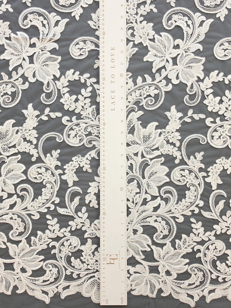 May include: Off-white lace fabric featuring an intricate floral and vine pattern with embroidered details. A white ruler labeled LACE TO LOVE is placed vertically over the fabric, showing measurements in both inches and centimeters.