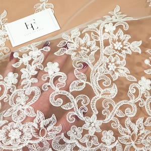 May include: White floral lace fabric with a delicate pattern. The lace is on a sheer mesh background. The fabric is perfect for adding a touch of elegance to any project.