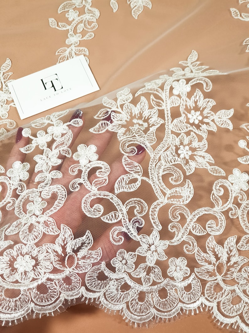 May include: White floral lace fabric with a delicate pattern. The lace is on a sheer mesh background. The fabric is perfect for adding a touch of elegance to any project.
