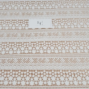 May include: White crochet lace fabric with a repeating pattern of flowers and geometric shapes. The fabric has a delicate and intricate design.