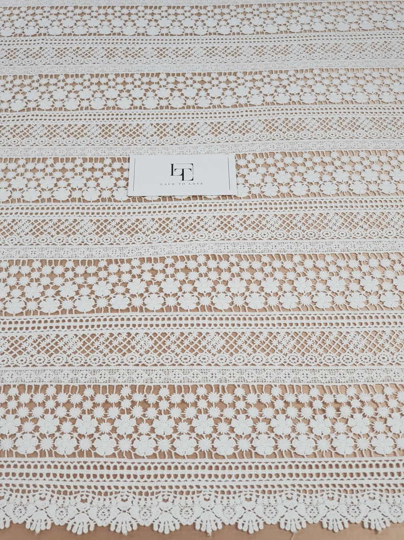 May include: White crochet lace fabric with a repeating pattern of flowers and geometric shapes. The fabric has a delicate and intricate design.