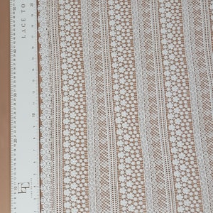 May include: White lace fabric with a repeating pattern of small flowers and geometric designs. The fabric has a delicate, airy feel and is perfect for adding a touch of elegance to any project.