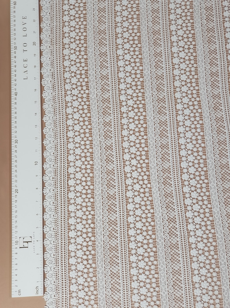 May include: White lace fabric with a repeating pattern of small flowers and geometric designs. The fabric has a delicate, airy feel and is perfect for adding a touch of elegance to any project.