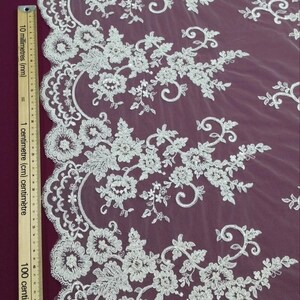 May include: Close-up of white floral lace fabric. The lace features detailed embroidered flowers and leaves, with a scalloped edge. The fabric is set against a deep purple background, creating a contrast that highlights the delicate design.