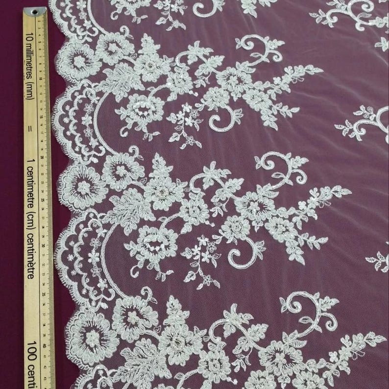 May include: Close-up of white floral lace fabric. The lace features detailed embroidered flowers and leaves, with a scalloped edge. The fabric is set against a deep purple background, creating a contrast that highlights the delicate design.