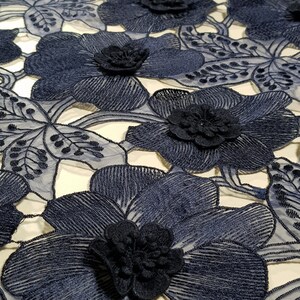 May include: A navy blue lace fabric featuring a repeating pattern of large, dimensional floral motifs. The flowers are constructed with intricate threadwork and raised, layered centres, connected by delicate, sheer mesh netting.