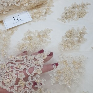 May include: Beige lace fabric with floral embroidery and beaded accents. The fabric features intricate designs and is partially visible with a hand underneath. A tag with the text "LACE TO LOVE" is also visible.