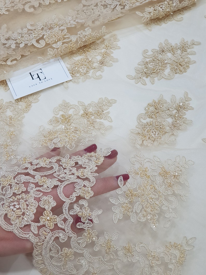 May include: Beige lace fabric with floral embroidery and beaded accents. The fabric features intricate designs and is partially visible with a hand underneath. A tag with the text "LACE TO LOVE" is also visible.