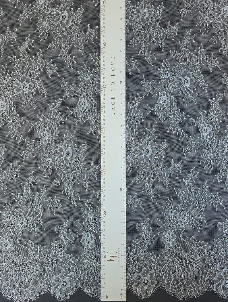 Pode incluir: Tecido de renda floral branca com um padr&atilde;o delicado. A renda apresenta um design repetido de flores e folhas. Uma r&eacute;gua com o texto "LACE TO LOVE" &eacute; colocada no tecido, com medidas em cm.