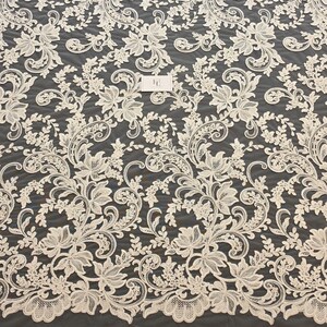 May include: An off-white lace fabric featuring an intricate floral and scrolling vine pattern with detailed embroidery. A small rectangular white tag with the letters FE is visible near the center of the textile, showcasing the delicate mesh base.