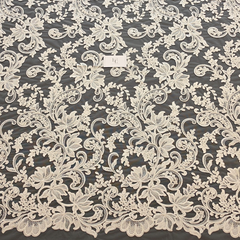 May include: An off-white lace fabric featuring an intricate floral and scrolling vine pattern with detailed embroidery. A small rectangular white tag with the letters FE is visible near the center of the textile, showcasing the delicate mesh base.