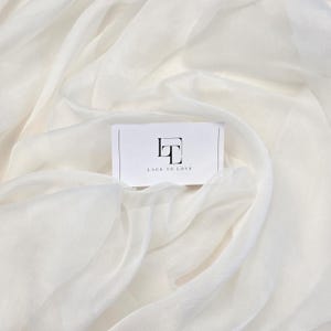 May include: White, flowing fabric with soft folds and a subtle texture. A business card with the text "LACE TO LOVE" is placed on top. The card features a minimalist logo in black.