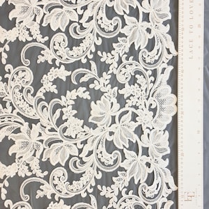 May include: Off-white lace fabric featuring an intricate floral and vine pattern with scalloped edges. A white ruler is positioned along the right side, showing measurements in both inches and centimeters, with the text LACE TO LOVE printed on it.