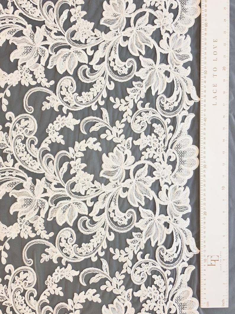May include: Off-white lace fabric featuring an intricate floral and vine pattern with scalloped edges. A white ruler is positioned along the right side, showing measurements in both inches and centimeters, with the text LACE TO LOVE printed on it.
