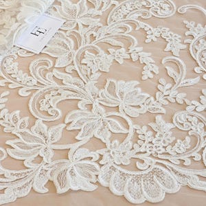 May include: White floral lace fabric featuring intricate embroidery and scalloped edges layered over a solid beige background. A small white rectangular tag with the black logo LT and the text LACE TO LOVE is placed in the upper left corner.