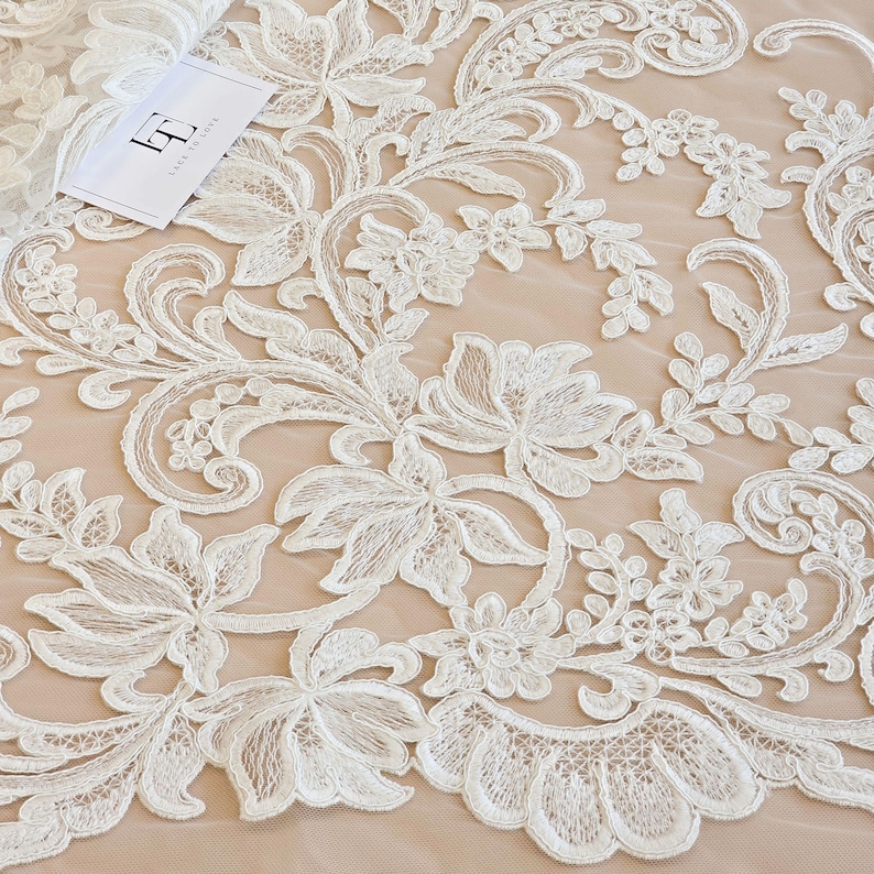 May include: White floral lace fabric featuring intricate embroidery and scalloped edges layered over a solid beige background. A small white rectangular tag with the black logo LT and the text LACE TO LOVE is placed in the upper left corner.