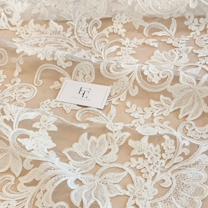 May include: White lace fabric featuring an intricate floral and paisley embroidery pattern on a sheer mesh base. A small rectangular white card with the black text LT LACE TO LOVE is placed on the fabric, showcasing the detailed needlework.