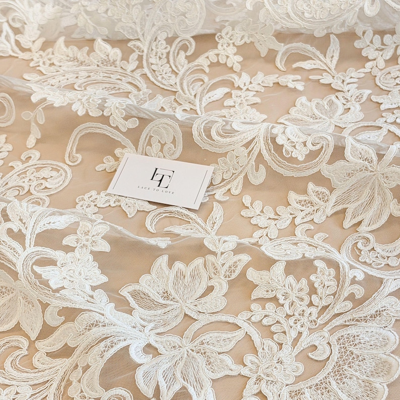 May include: White lace fabric featuring an intricate floral and paisley embroidery pattern on a sheer mesh base. A small rectangular white card with the black text LT LACE TO LOVE is placed on the fabric, showcasing the detailed needlework.