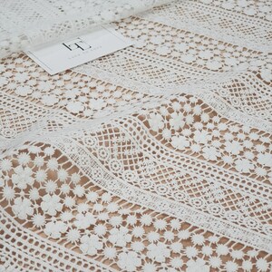 May include: White crochet lace fabric with a delicate floral pattern. The lace has a vertical striped design with different sized flowers and geometric shapes.