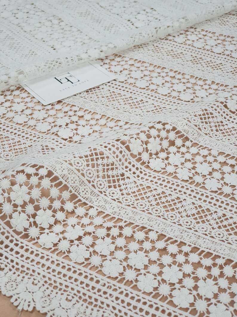 May include: White crochet lace fabric with a delicate floral pattern. The lace has a vertical striped design with different sized flowers and geometric shapes.
