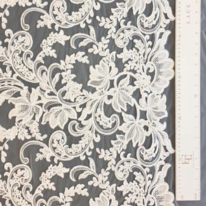 May include: Off-white lace fabric featuring an intricate floral and scrolling vine pattern with detailed embroidery. A white ruler is positioned along the right edge, showing measurements in centimeters and inches up to 48 centimeters.