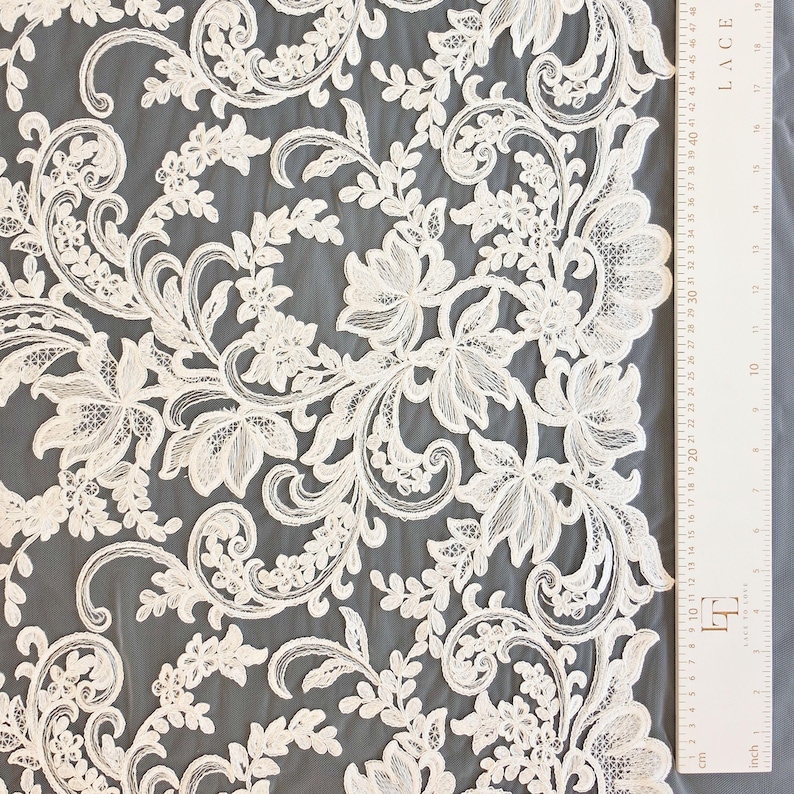 May include: Off-white lace fabric featuring an intricate floral and scrolling vine pattern with detailed embroidery. A white ruler is positioned along the right edge, showing measurements in centimeters and inches up to 48 centimeters.