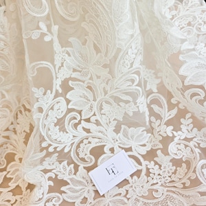 May include: Off-white lace fabric featuring an intricate floral and paisley embroidery pattern on a sheer mesh base. A small white rectangular tag with the black logo LT and the text LACE TO LOVE is placed on the surface of the fabric.