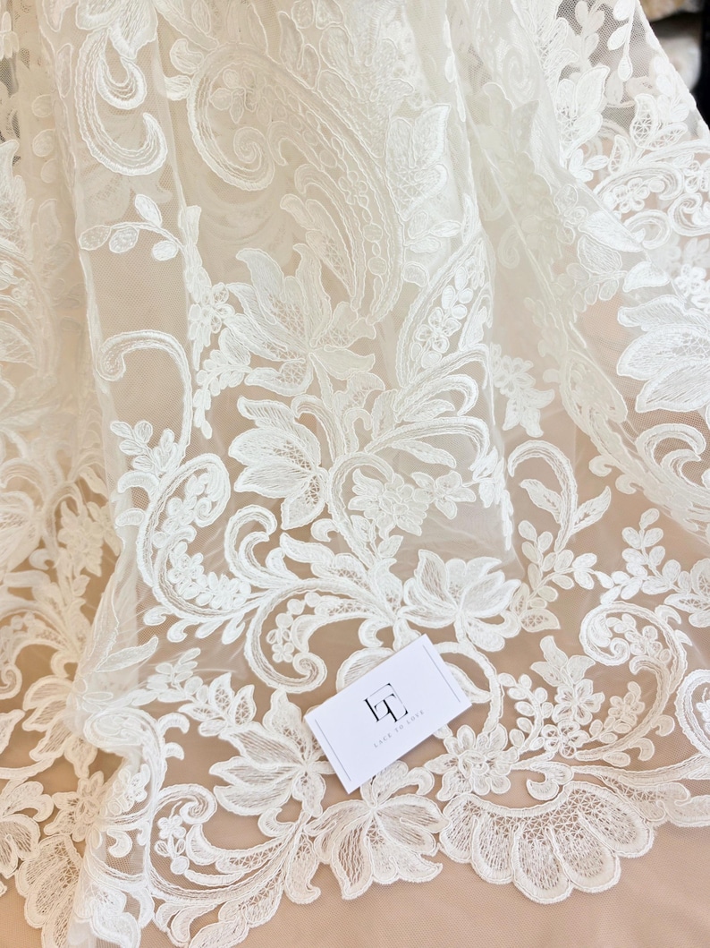 May include: Off-white lace fabric featuring an intricate floral and paisley embroidery pattern on a sheer mesh base. A small white rectangular tag with the black logo LT and the text LACE TO LOVE is placed on the surface of the fabric.