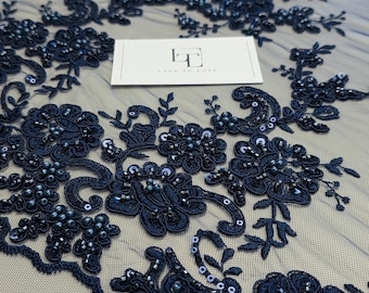 May include: Navy blue lace fabric featuring intricate floral embroidery, dark beads, and sequins on a sheer mesh base. A white rectangular card with the logo LT and the text LACE TO LOVE is placed on the fabric surface.