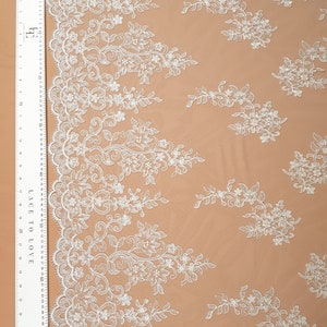 May include: White floral lace fabric with a delicate pattern. The lace has a scalloped edge and is perfect for crafting, sewing, or decorating.