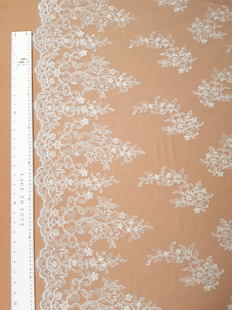May include: White floral lace fabric with a delicate pattern. The lace has a scalloped edge and is perfect for crafting, sewing, or decorating.