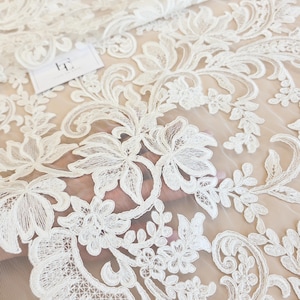 May include: A sheer white lace fabric featuring an intricate floral and vine embroidery pattern. The design consists of detailed leaves and blossoms stitched onto a fine mesh base. A small white rectangular tag with black text is visible.