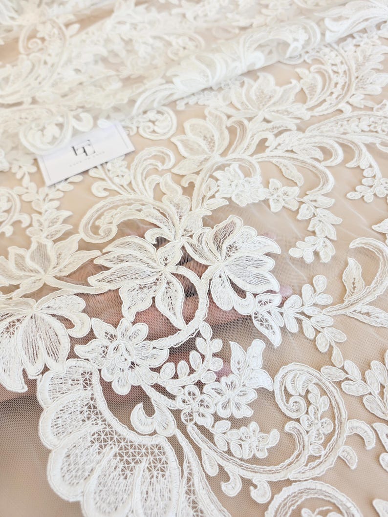May include: A sheer white lace fabric featuring an intricate floral and vine embroidery pattern. The design consists of detailed leaves and blossoms stitched onto a fine mesh base. A small white rectangular tag with black text is visible.