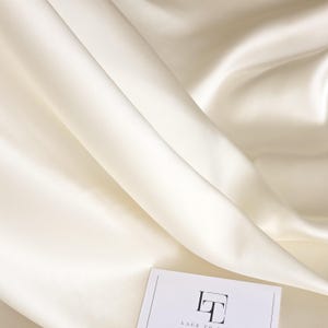 Könnte beinhalten: Elfenbeinfarbener Satin-Stoff mit weichen Falten und dezentem Glanz. Der Stoff ist drapiert und erzeugt eine fließende Textur. Eine weiße Karte mit einem Logo ist in der unteren rechten Ecke sichtbar. Der Stoff wirkt glatt und luxuriös.