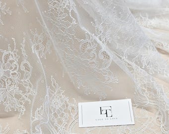 Puede incluir: Primer plano de tela de encaje blanco con patrones florales. El delicado encaje está drapeado, mostrando los intrincados diseños. Una pequeña tarjeta blanca con el texto "LACE TO LOVE" está en la esquina inferior derecha. La tela es ideal para ropa de novia u otras ocasiones especiales.