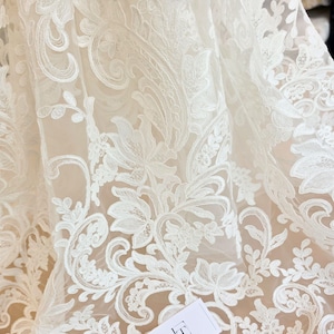 May include: A sheer off-white fabric featuring intricate white floral embroidery with swirling vine patterns. The delicate lace material displays a dense, layered botanical design with detailed stitching throughout the translucent mesh textile.