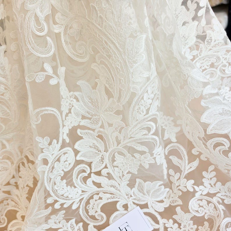 May include: A sheer off-white fabric featuring intricate white floral embroidery with swirling vine patterns. The delicate lace material displays a dense, layered botanical design with detailed stitching throughout the translucent mesh textile.